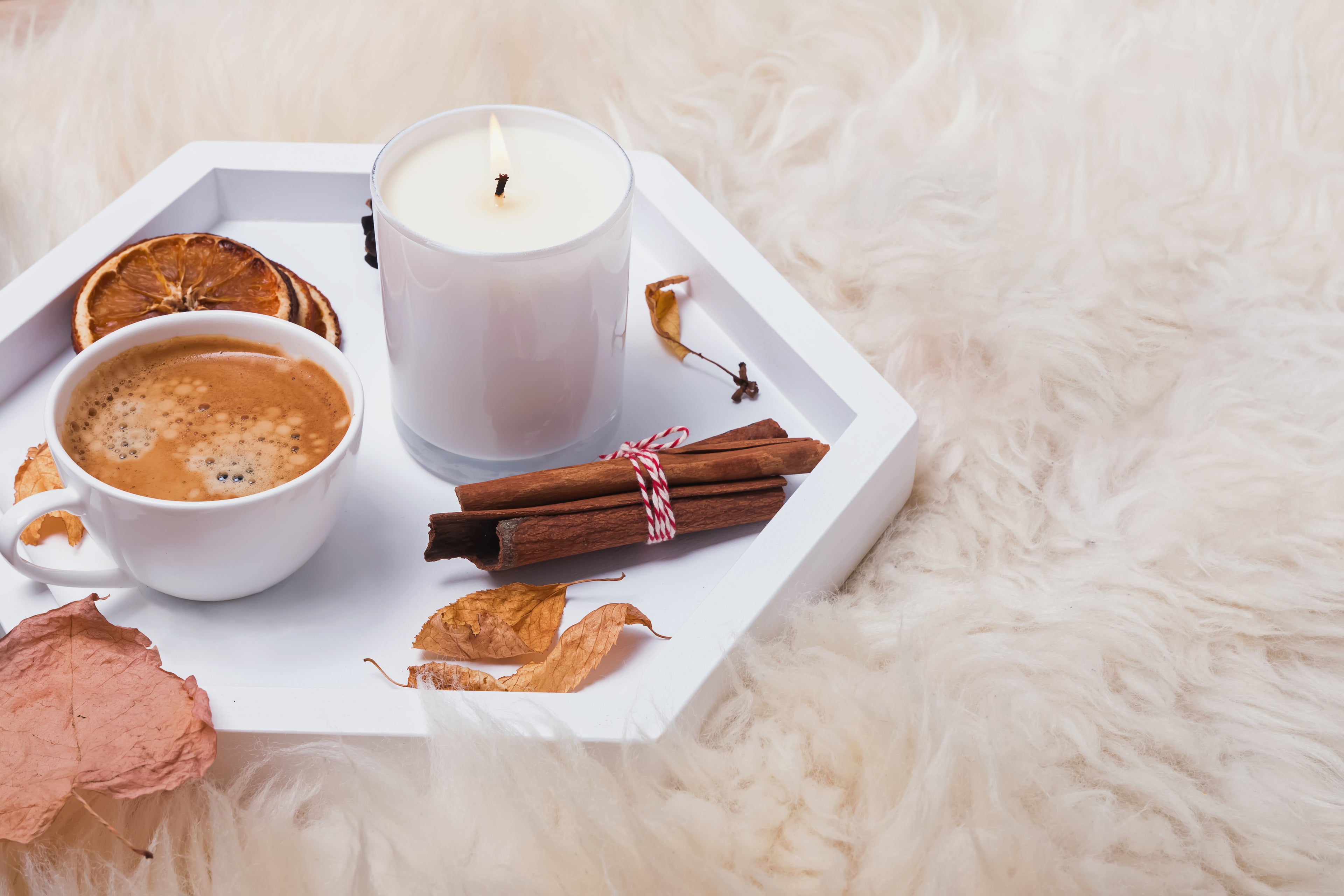 A tray with a burning candle, a hot coffee mug, cinnamon sticks, and some dried oranges laid on a plush shag white rug